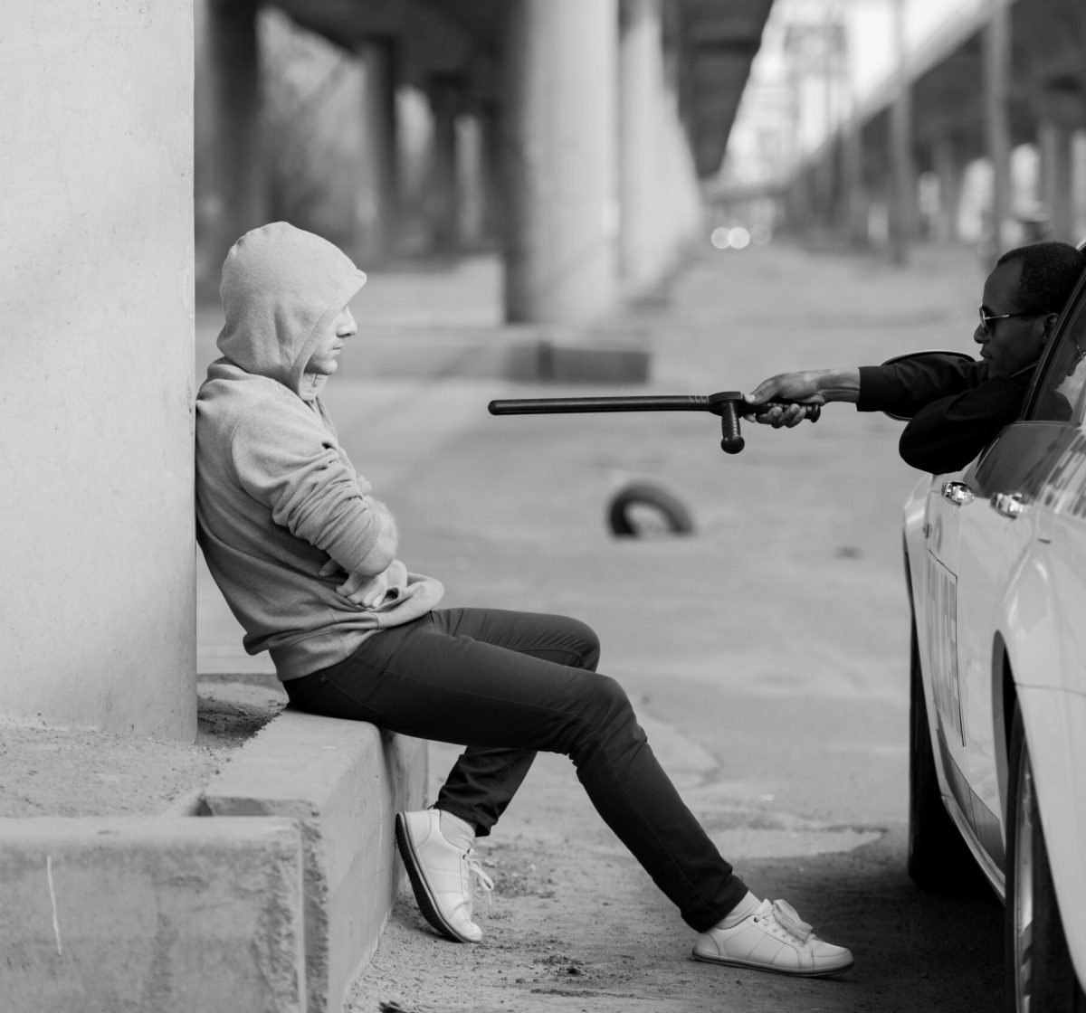 police officer pointing at hooded man with police bat while he sitting on street